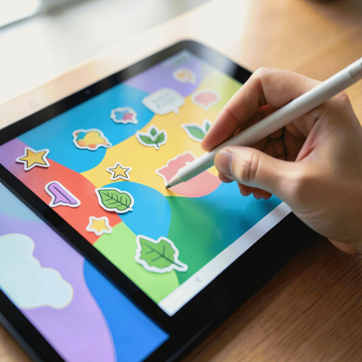 Tangan sedang menempelkan stiker digital yang berwarna-warni dan estetik di layar tablet, menunjukkan proses desain konten secara langsung.