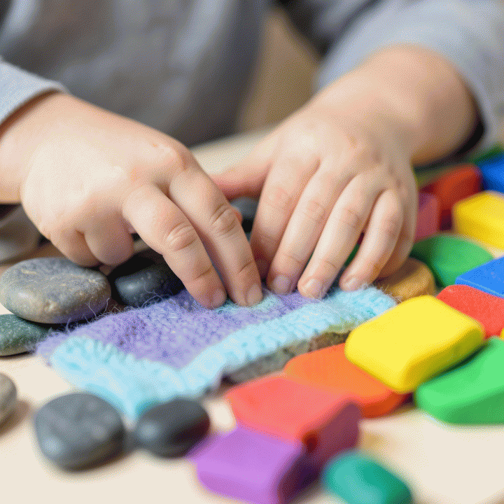 Close-up of a child&rsquo;s hands exploring tactile learning materials like stones, fabric, and playdough for sensory development.